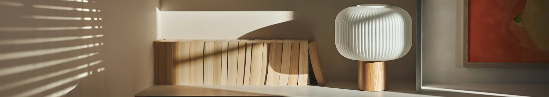 Una moderna lampada bianca a coste con base in legno si trova accanto a una fila di blocchi di legno chiaro, con la luce del sole che proietta ombre orizzontali.