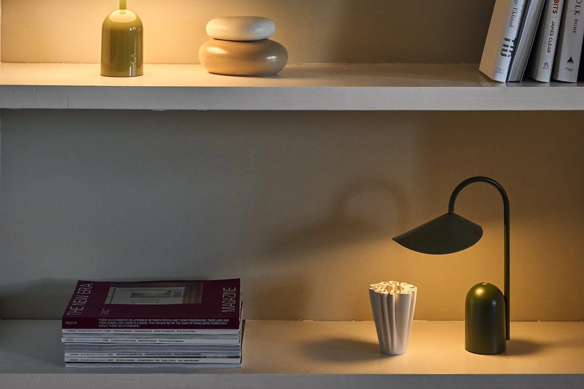 Tre mensole espongono diverse lampade da tavolo verdi e viola, insieme a libri, riviste e oggetti decorativi.