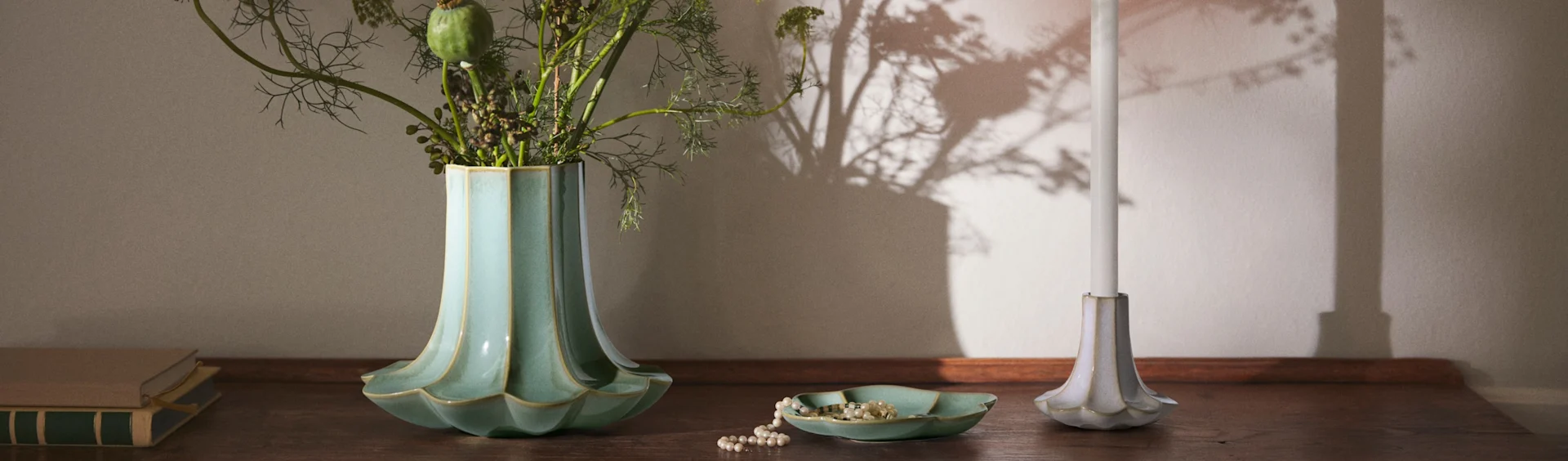 Un vaso scanalato color verde acqua con fogliame, una collana di perle in un piatto abbinato, una candela bianca in un portacandele scanalato e libri su una superficie di legno.