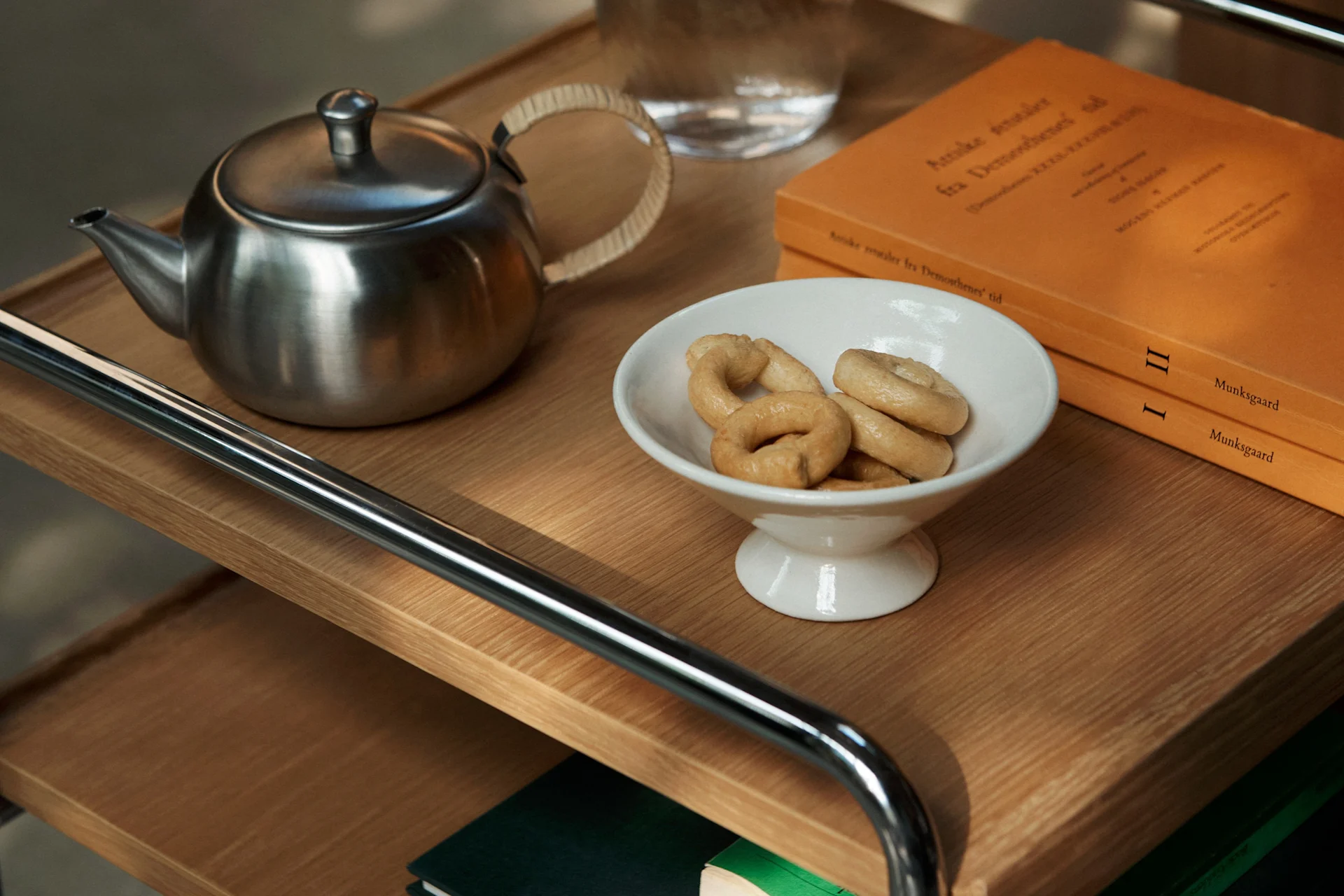 Una teiera in acciaio inossidabile, una ciotola bianca con biscotti ad anello e due libri arancioni su uno scaffale di legno.