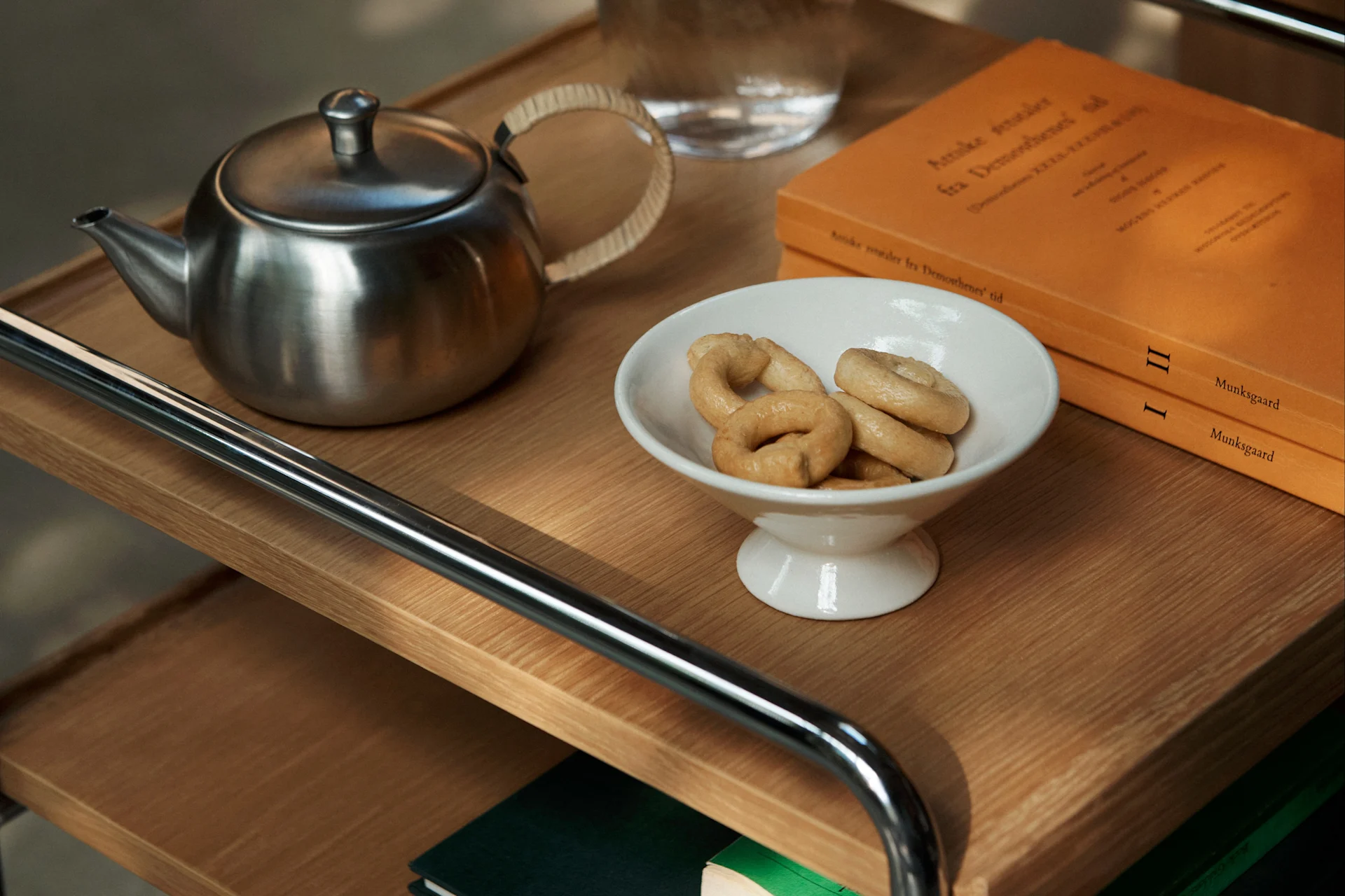 Una teiera in metallo, una ciotola bianca di biscotti ad anello e due libri arancioni su un carrello di legno.