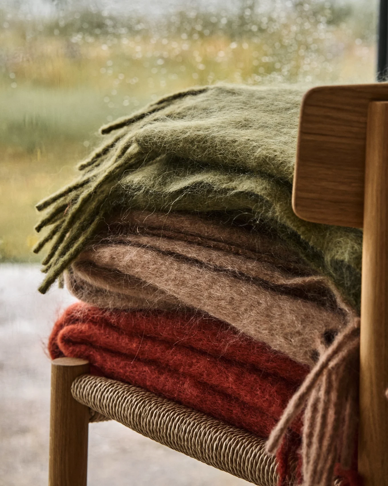 Pila di tre morbide coperte verde oliva, beige e rosso-arancio su una sedia di legno, con una finestra piovosa sullo sfondo.