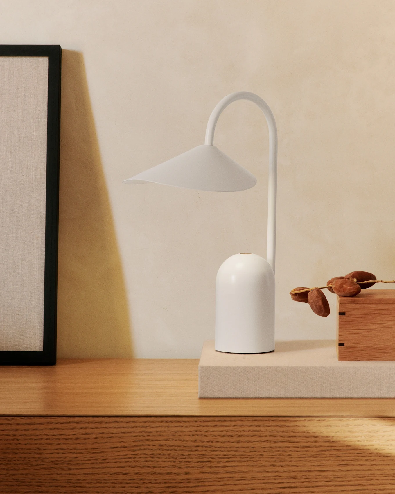 Una lampada da tavolo bianca minimalista con braccio curvo su una cassettiera di legno, accanto a un quadro incorniciato e una scatola di legno con datteri.