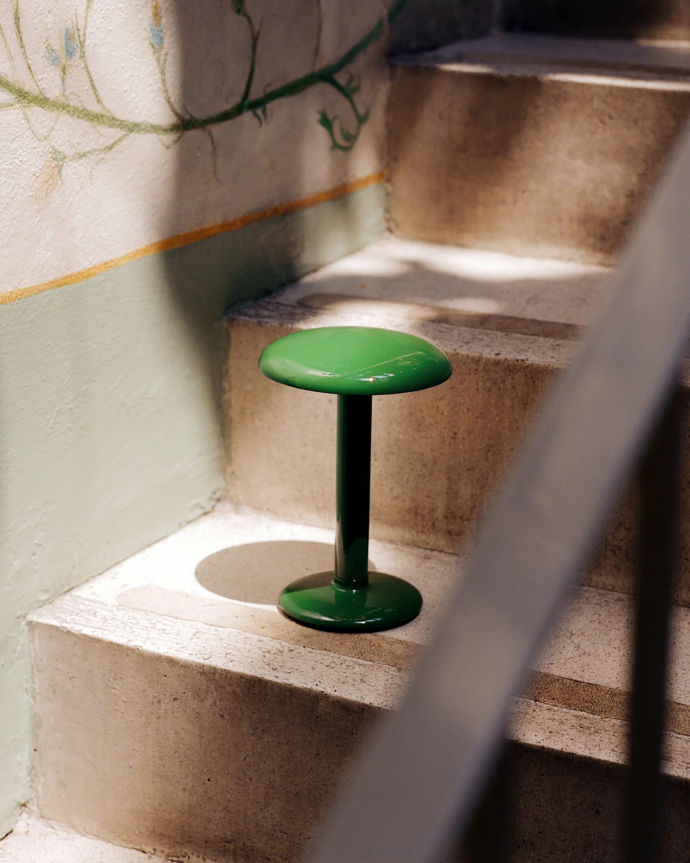 Uno sgabello verde lucido e brillante con piano e base rotondi si trova su una scala di cemento accanto a una parete con un murale floreale sbiadito.