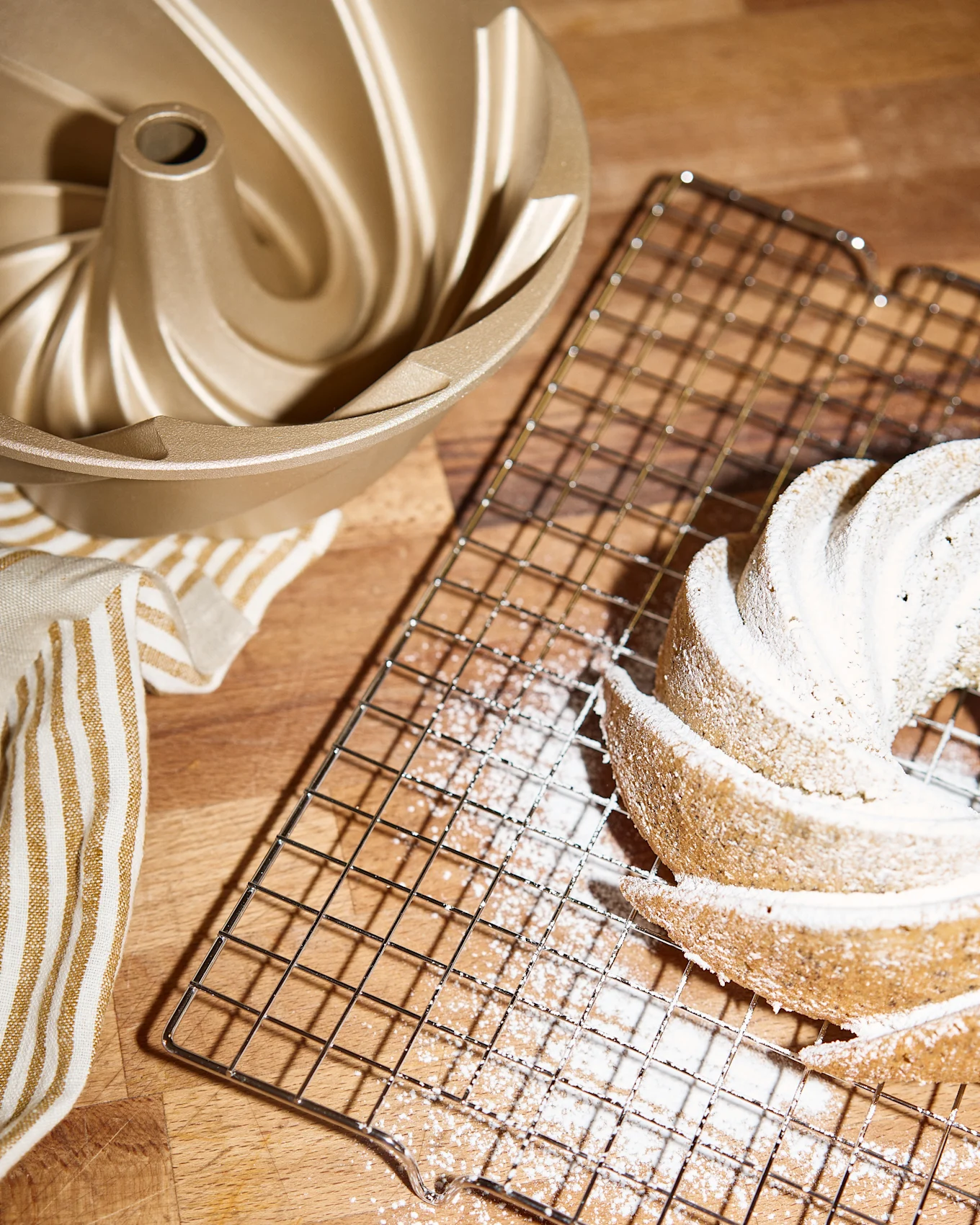 Uno stampo dorato per bundt cake, una griglia di raffreddamento con una bundt cake spolverata di zucchero a velo e un asciugamano a righe su una superficie di legno.
