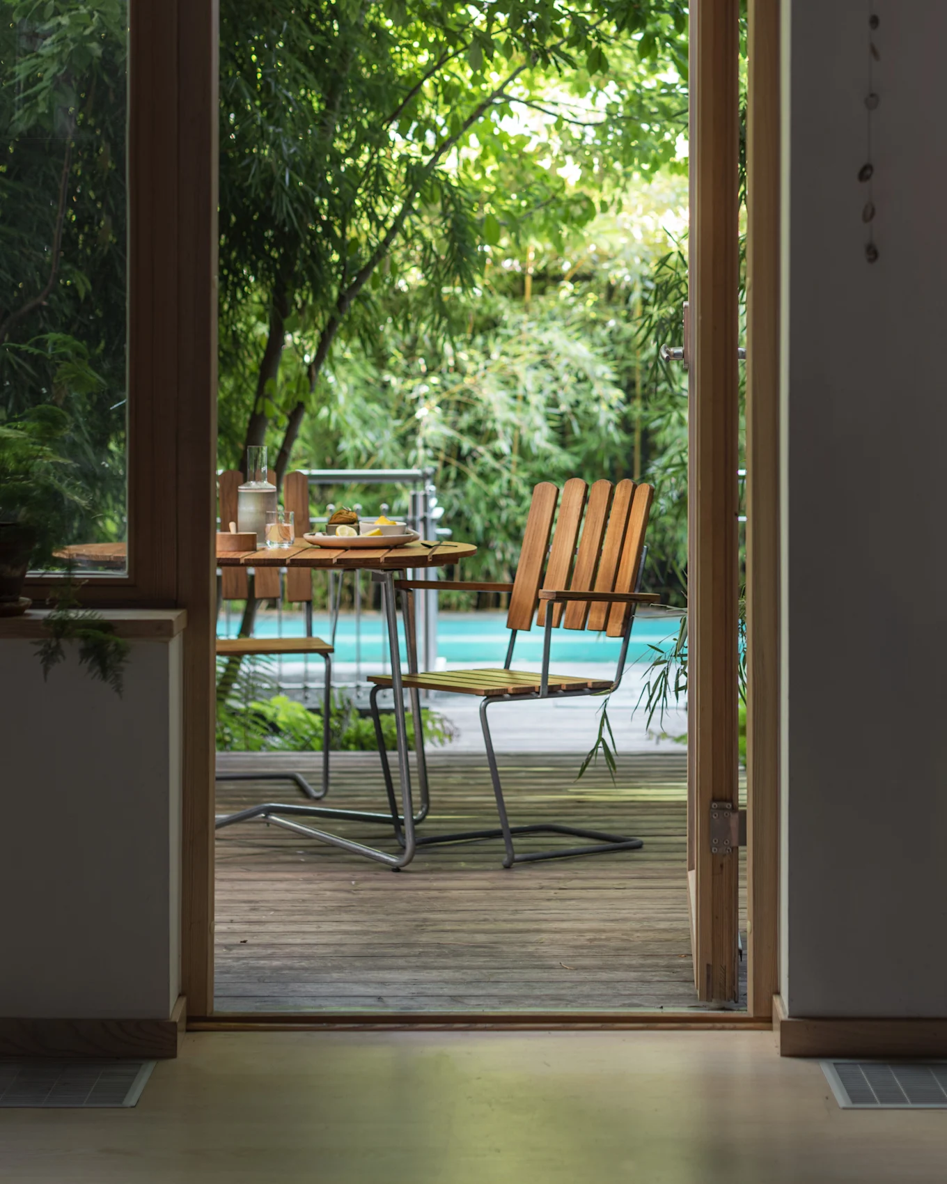 Vista attraverso una porta aperta su un patio esterno con un tavolo in legno, sedie e una piscina circondata da piante verdi.