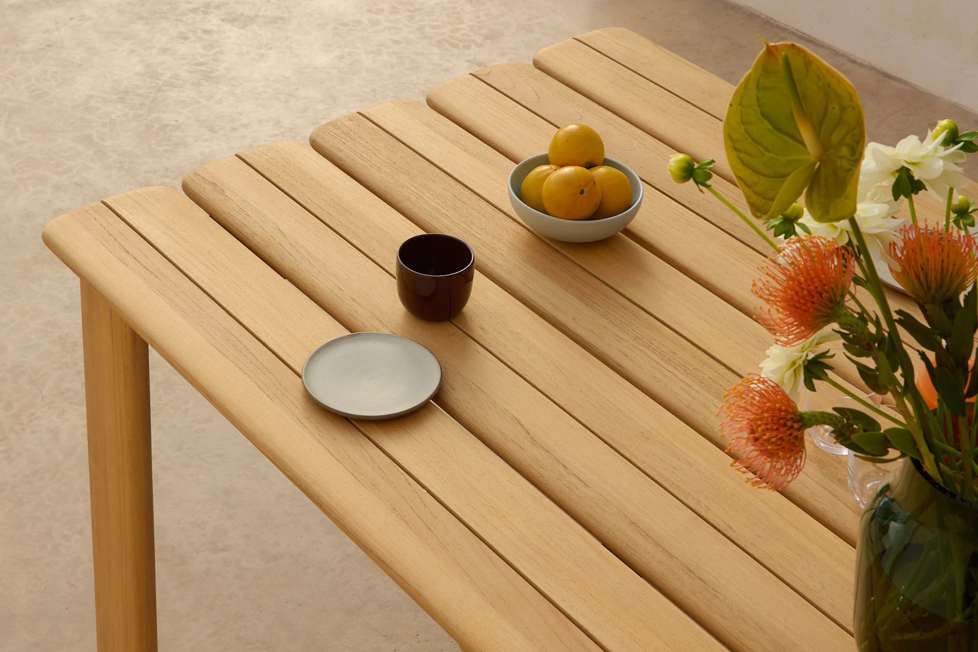 Un tavolo in legno chiaro a doghe con una ciotola di frutti gialli, una tazza scura, un piatto grigio e un vaso di fiori arancioni e bianchi.