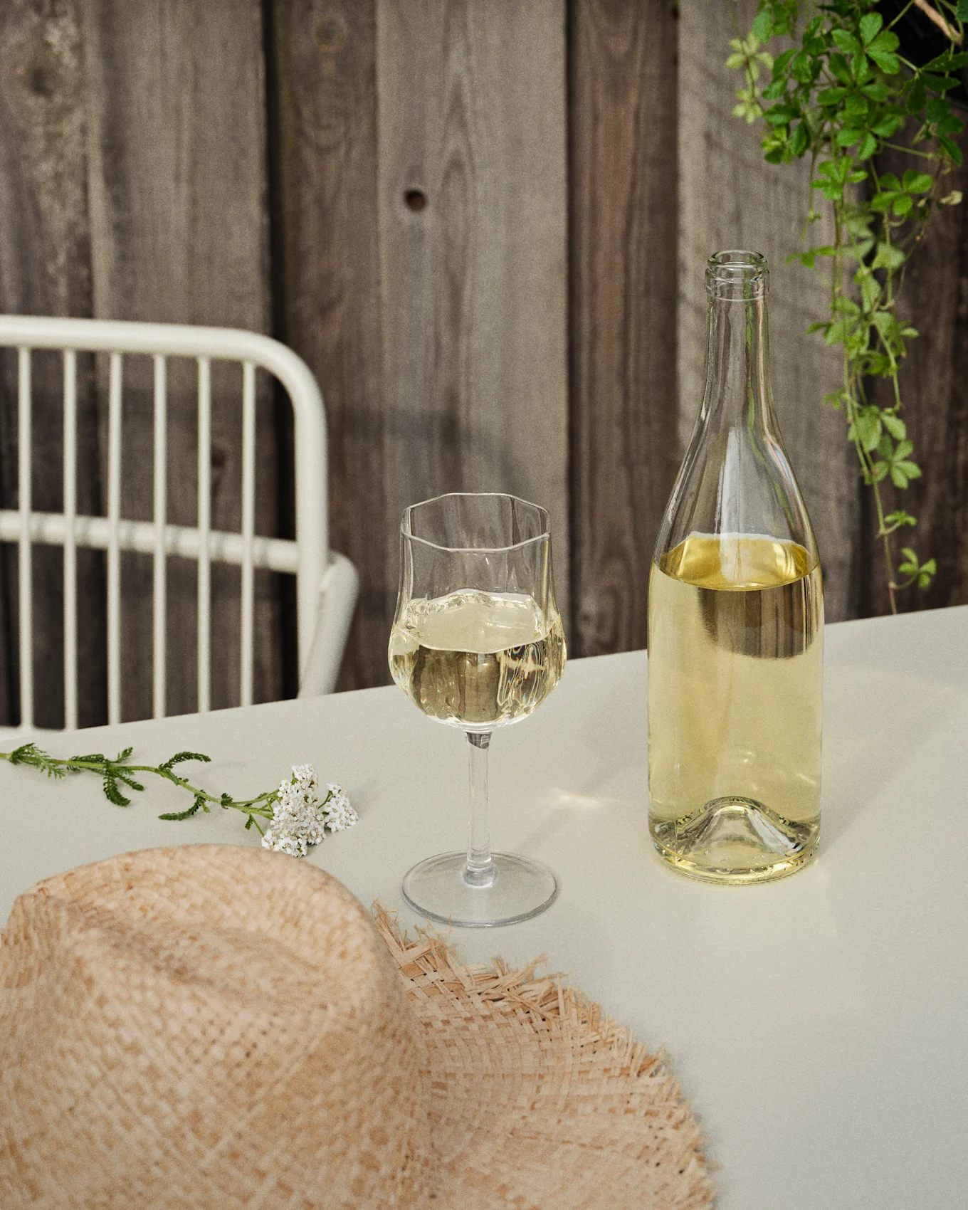 Una bottiglia e un bicchiere di vino bianco su un tavolo con un cappello di paglia e fiori bianchi, contro una parete di legno e viti verdi.