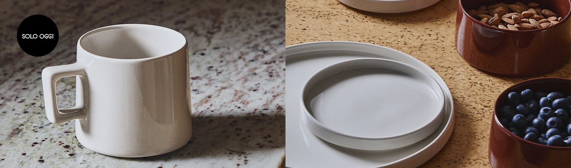 Una tazza bianca e rotonda con manico squadrato appoggiata su un piano di granito. Accanto, piatti bianchi impilati e ciotole marroni con mandorle e mirtilli su una superficie di sughero.