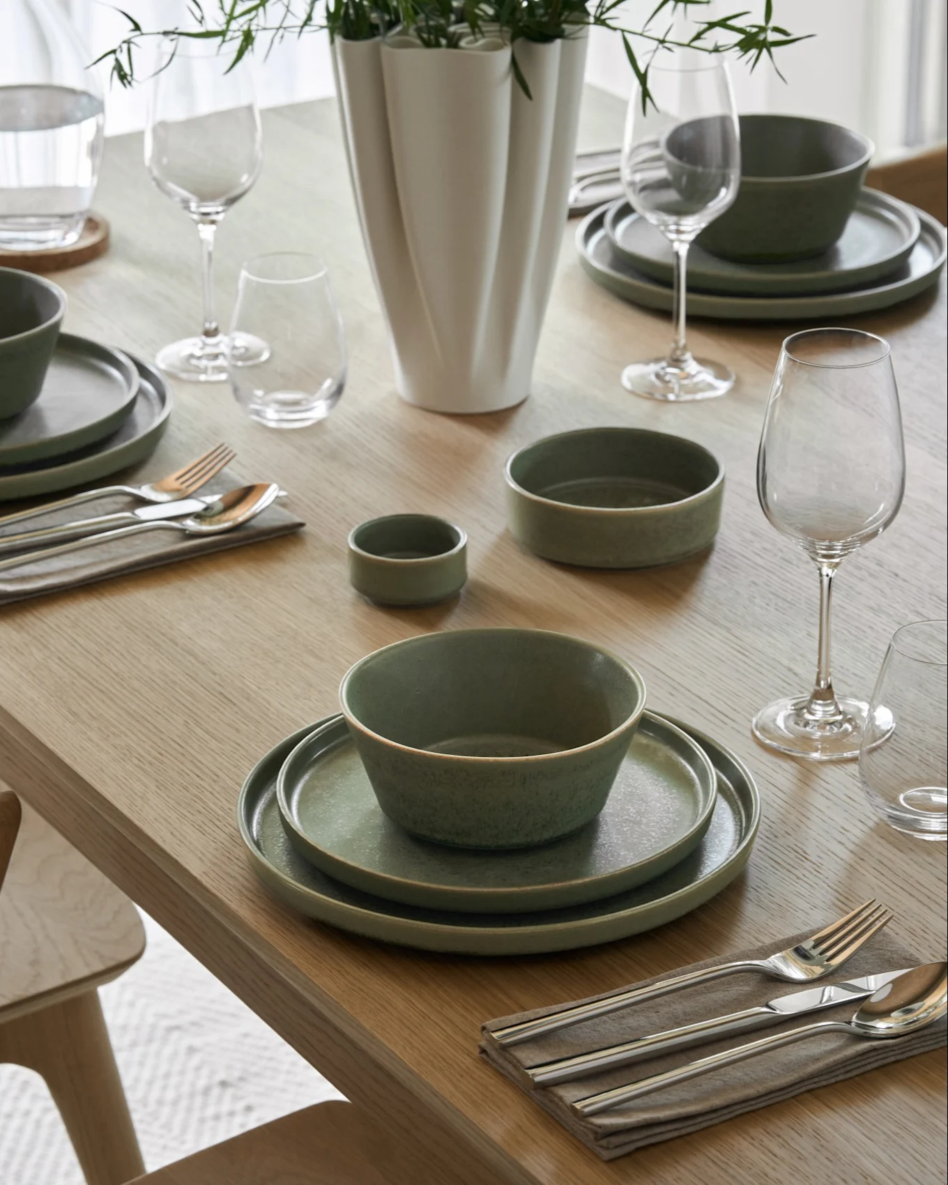 Tavolo da pranzo apparecchiato con ciotole di zuppa cremosa, piatti abbinati, bicchieri da vino e acqua, posate e un vaso con fogliame verde.