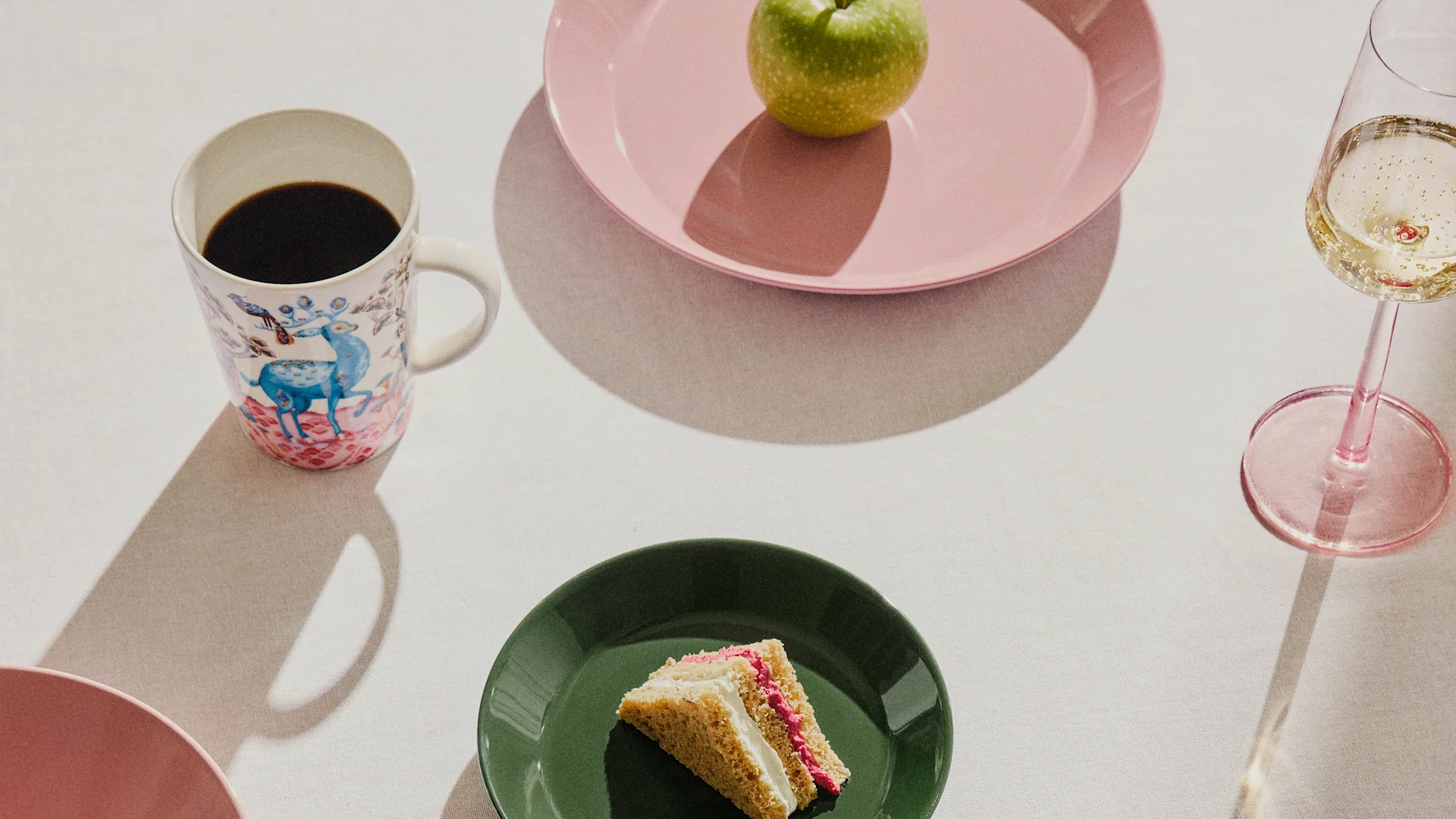 Tavola apparecchiata con una tazza da caffè a fantasia, una mela verde su un piatto rosa, una fetta di torta su un piatto verde e un bicchiere di champagne.