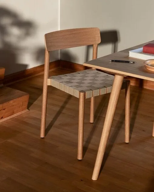 Una sedia in legno chiaro con seduta a scacchi intrecciata accanto a un tavolo con libri e una penna.