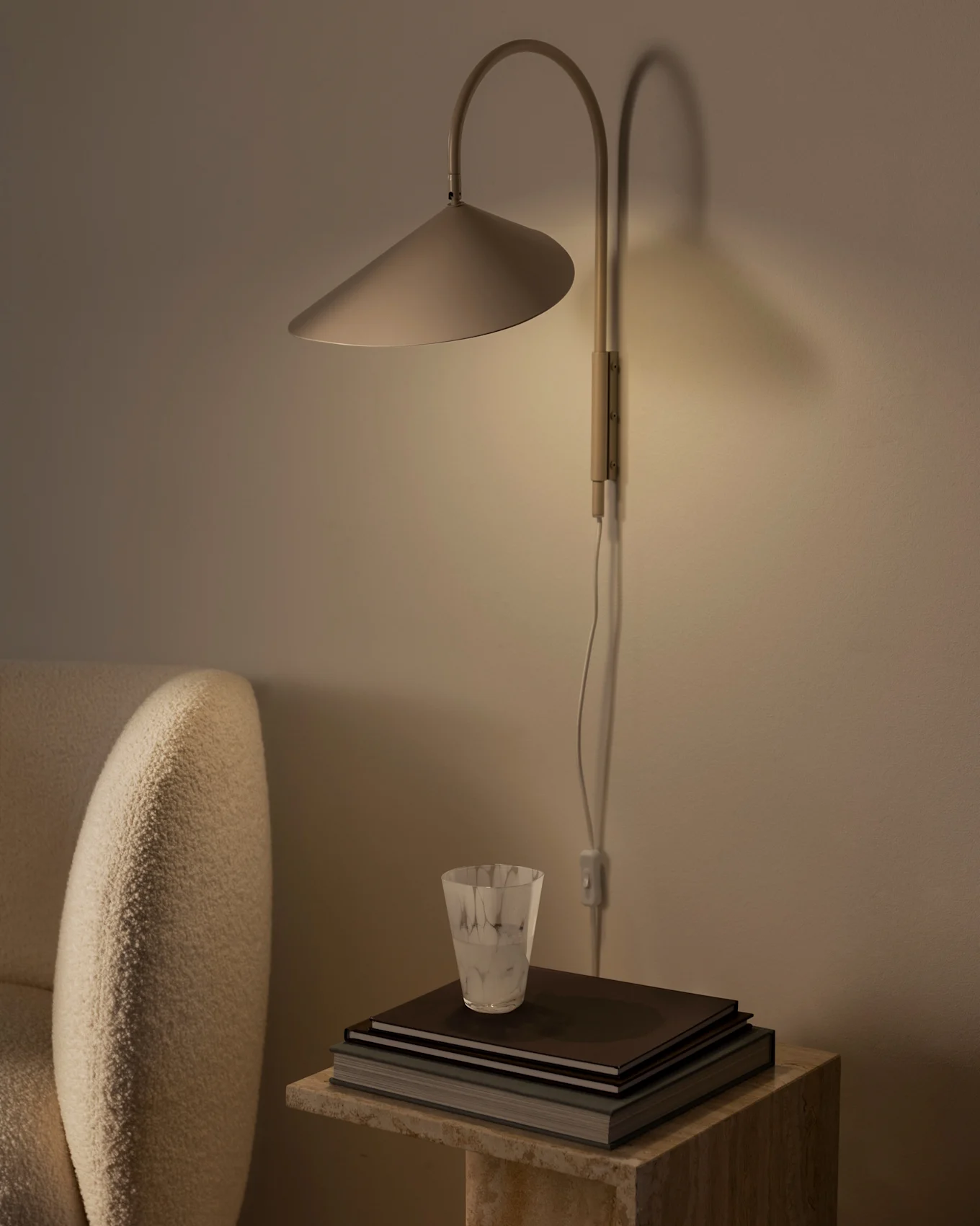 Lampada da parete illuminata con paralume conico sopra libri e bicchiere testurizzato su tavolo in pietra, accanto a una poltrona beige.