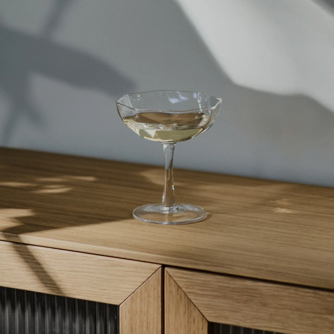 Un bicchiere coupe trasparente con una bevanda chiara su un mobile di legno chiaro, con ombre sulla parete.
