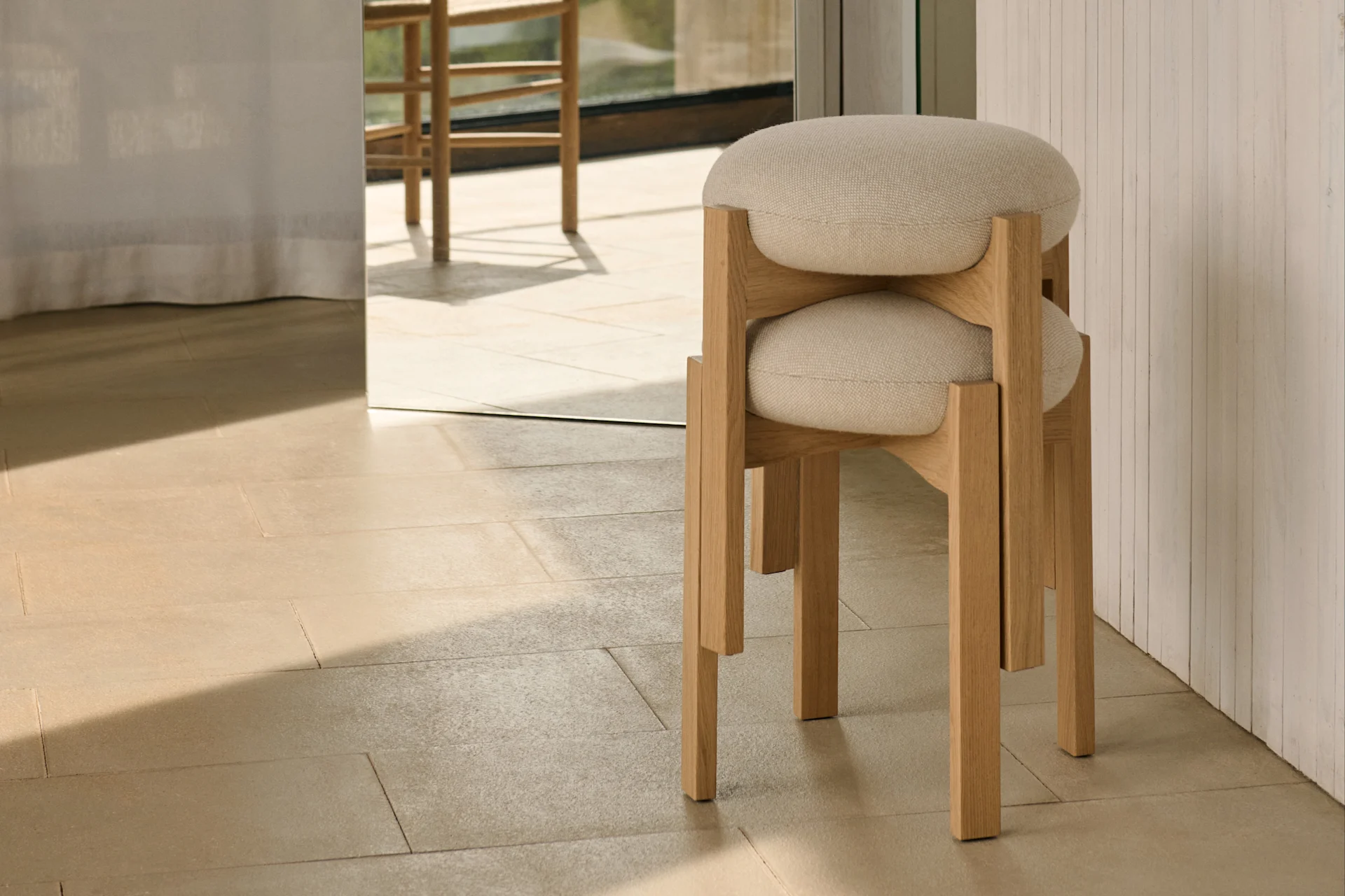 Due sgabelli minimalisti in legno chiaro con sedili rotondi imbottiti sono impilati su piastrelle chiare, con vista esterna soleggiata attraverso una grande finestra.