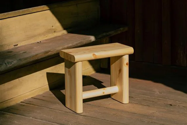Uno sgabello di legno chiaro con bordi arrotondati su un pavimento in legno, parzialmente illuminato dal sole.