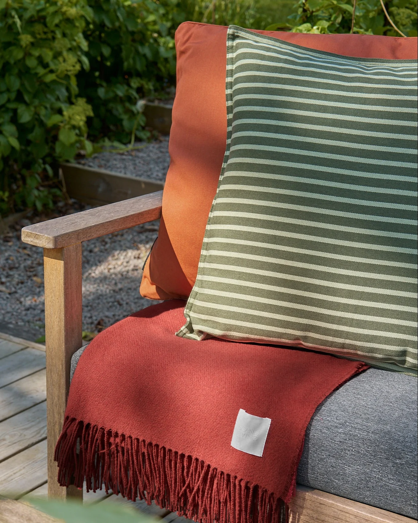 Sedia da esterno in legno con cuscino arancione, cuscino a righe verdi e bianche e coperta sfrangiata color terracotta.