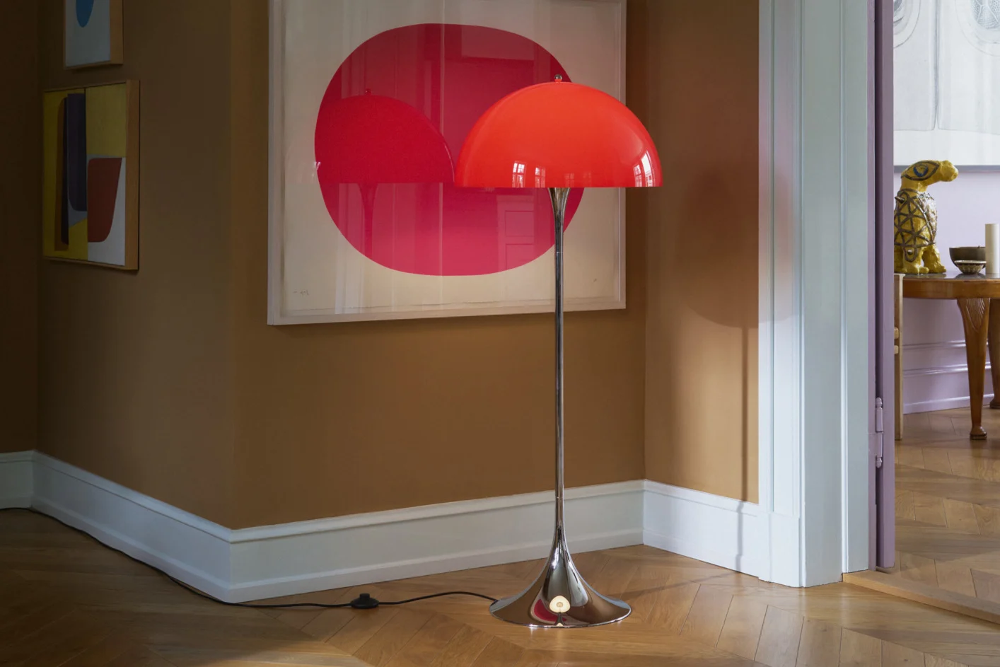 Una lampada da terra rossa brillante a cupola con stelo cromato su un pavimento in legno in una stanza con pareti marroni e arte moderna.