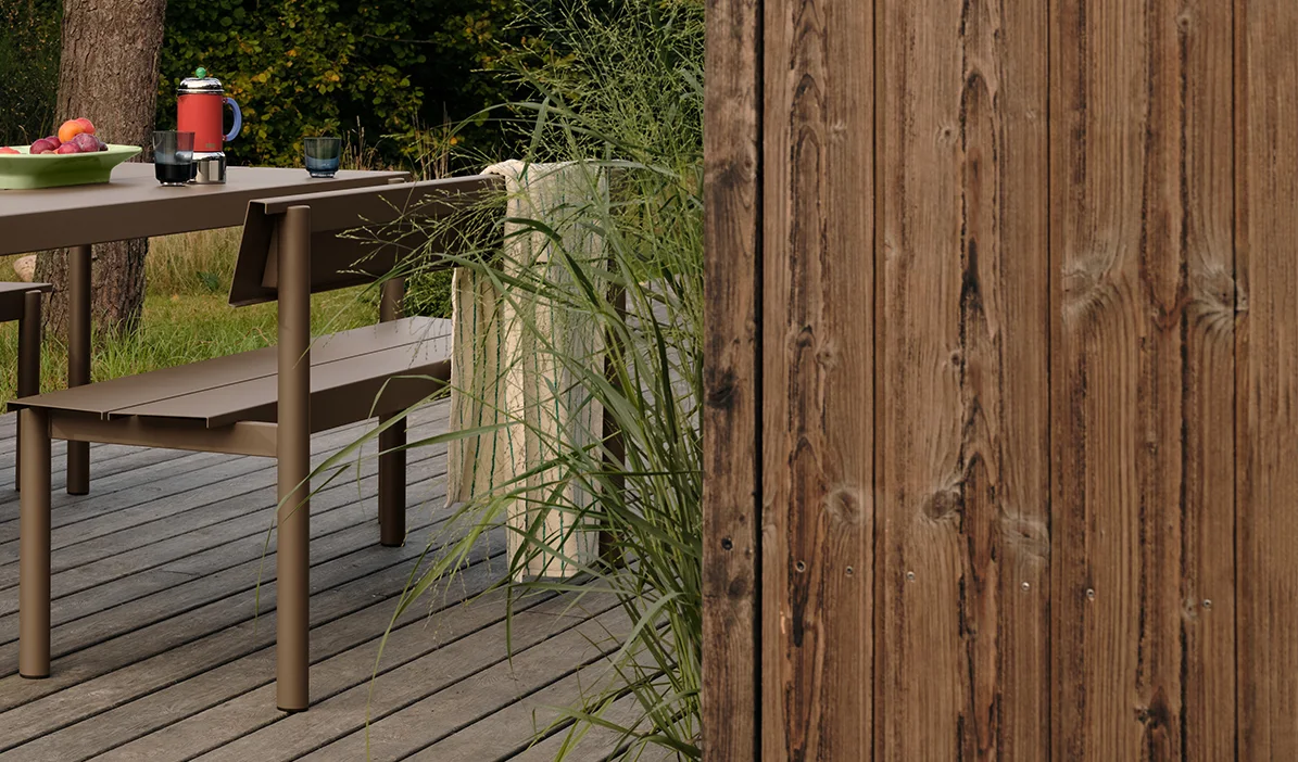 Tavolo e panca da esterno in metallo marrone su un ponte di legno con frutta e bevande, accanto a erba alta e una recinzione di legno.