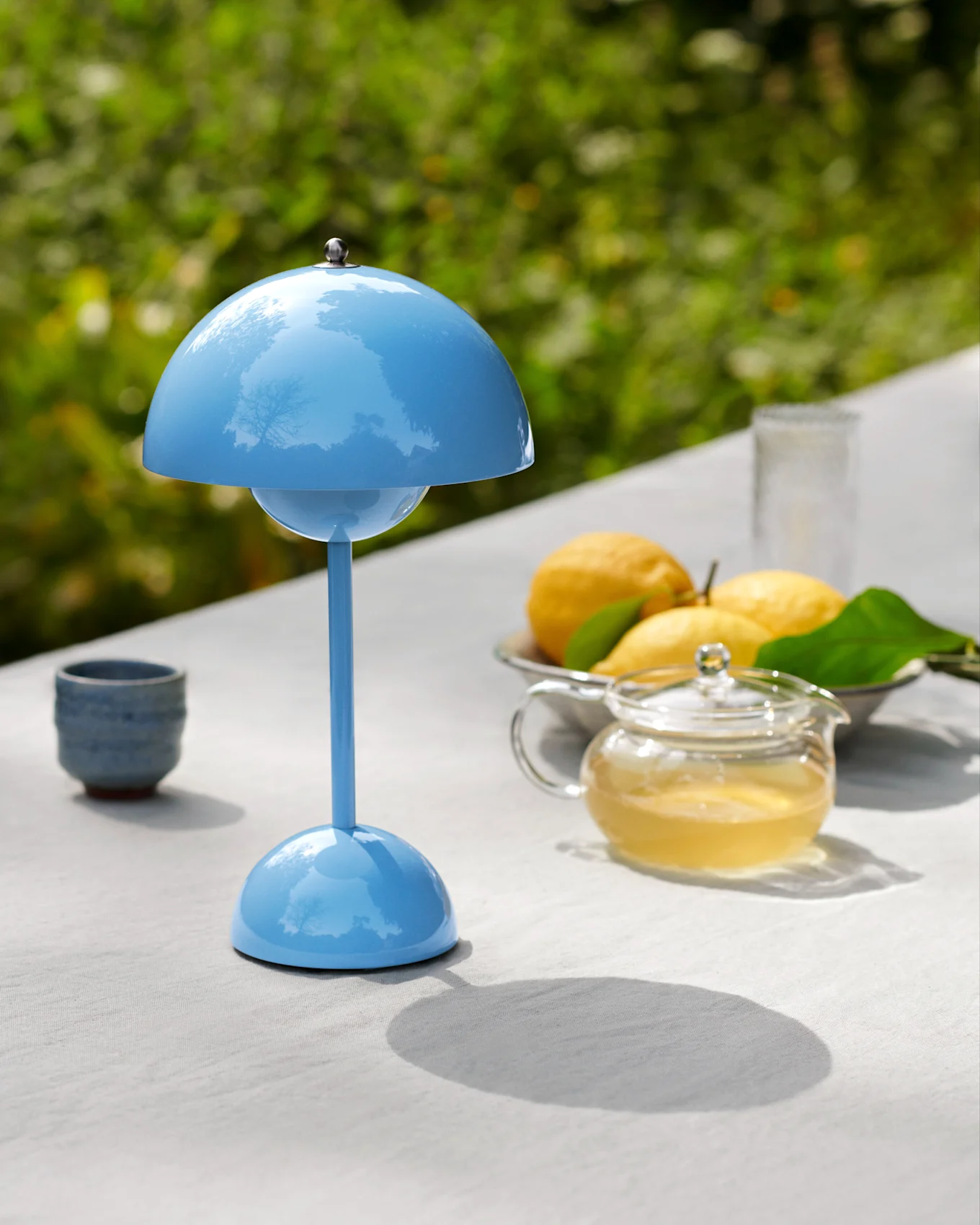 Una lampada da tavolo blu a forma di fungo su un tavolo da esterno con una teiera, limoni e una tazza.